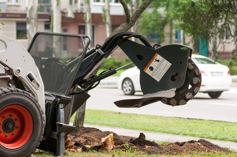 image of a brand new stump removal machine on a skid steer about to grind away a tree that's been cut down near a road with a car passing by